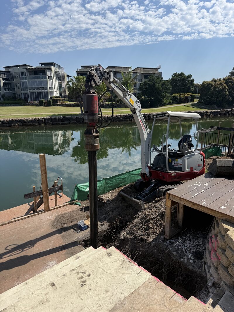 Mini excavator with drive head installing a screw pile for a waterfront home extension at Twin Waters